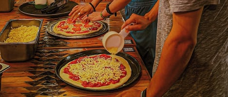 Um grupo de pessoas preparando pizzas em uma cozinha com mesas de madeira e ambiente rústico.