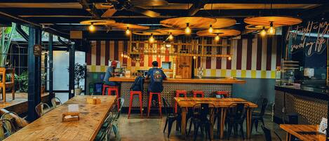 A restaurant interior with wooden tables, red stools, hanging light fixtures, and a bar area.