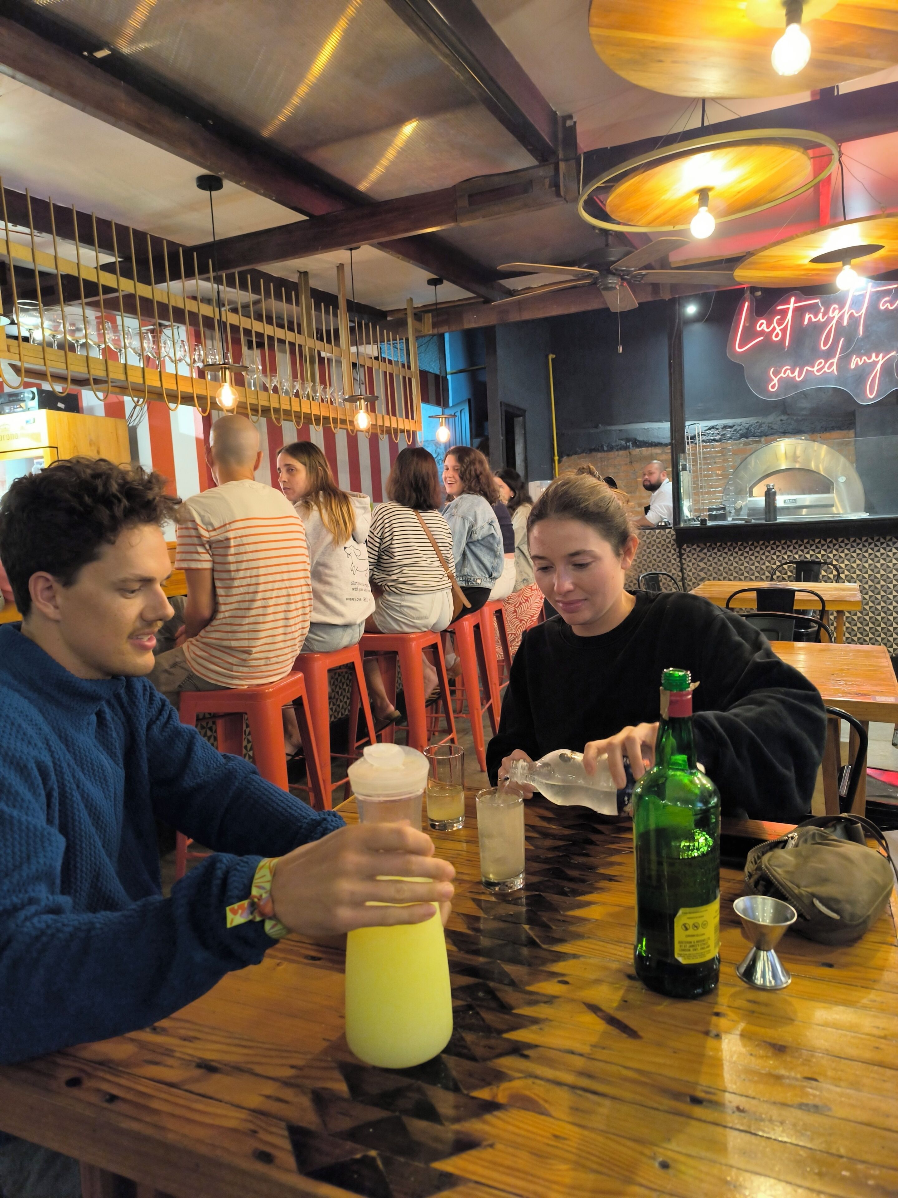A bar setting with patrons seated at tables, a neon sign, and a visible bar counter.