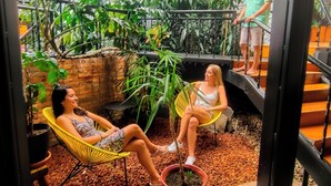 An indoor space with a high ceiling, metal beams, and a variety of potted plants. There are two people seated on chairs, one on a yellow chair and one on a white chair. A person is standing on a staircase. - Viajero San José Hostel (San José)