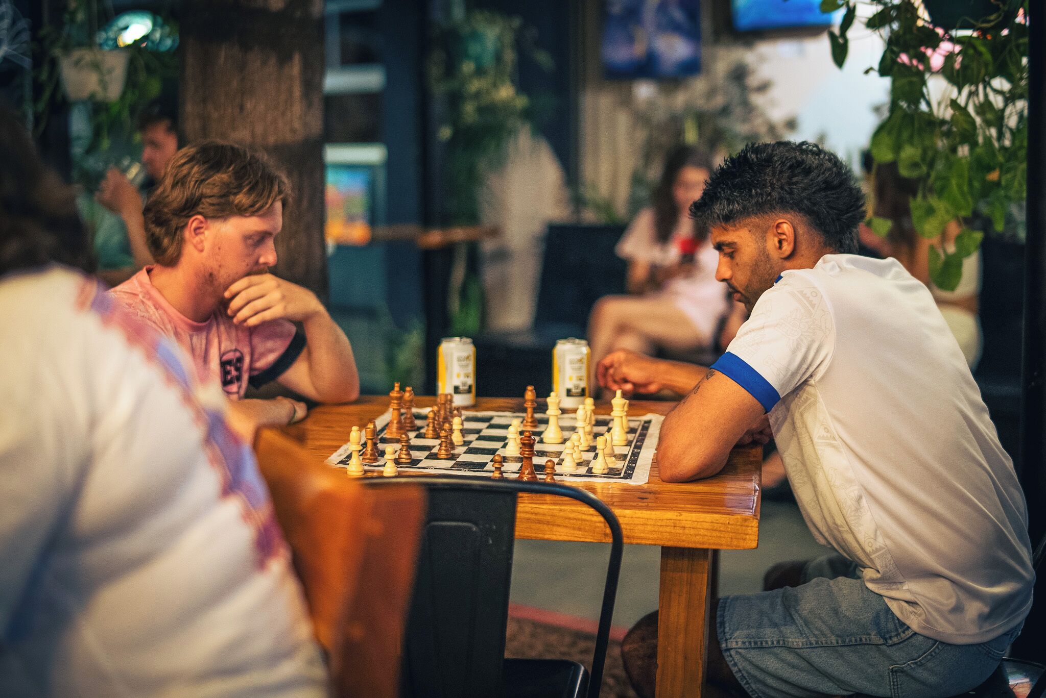 Two individuals playing chess at a wooden table in a room with plants and a television in the background.