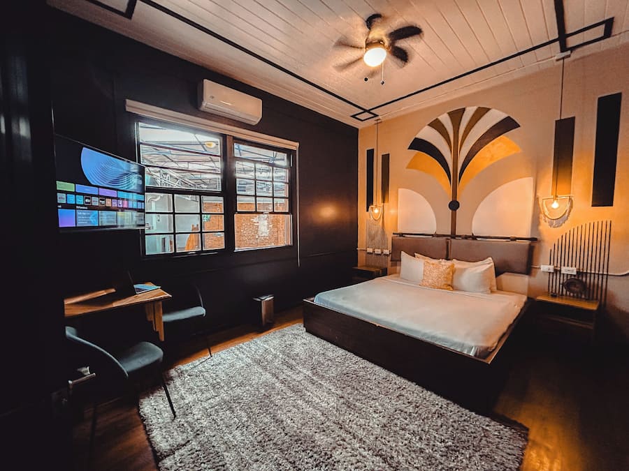 A bedroom with a bed, a desk, a chair, a TV, and a window with a view of buildings.