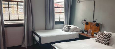 A hotel room with two single beds, a wooden nightstand, a desk with a lamp, and a window with curtains.