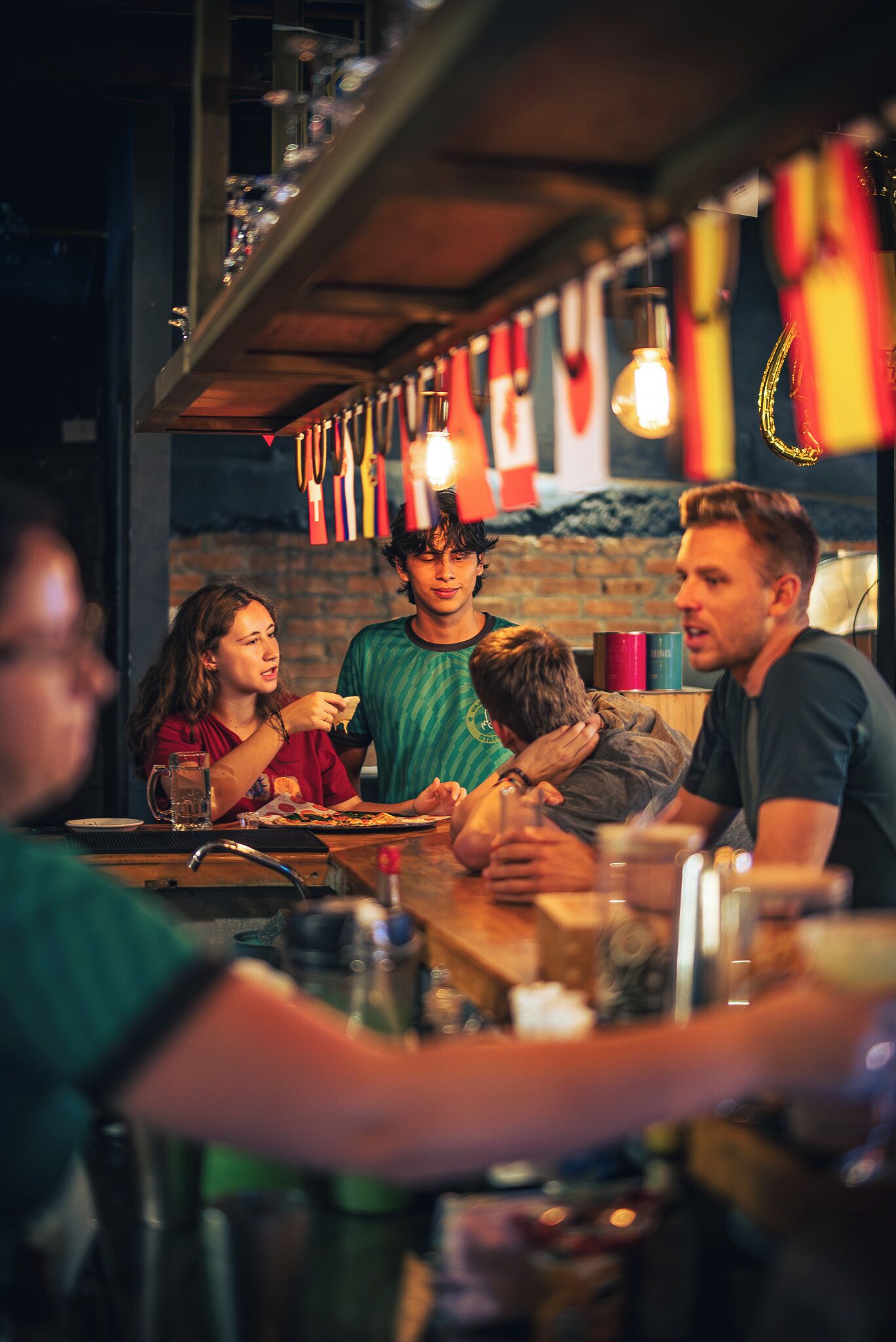 Un grupo de personas cenando en un bar con una pared de ladrillo y banderas colgando al fondo.