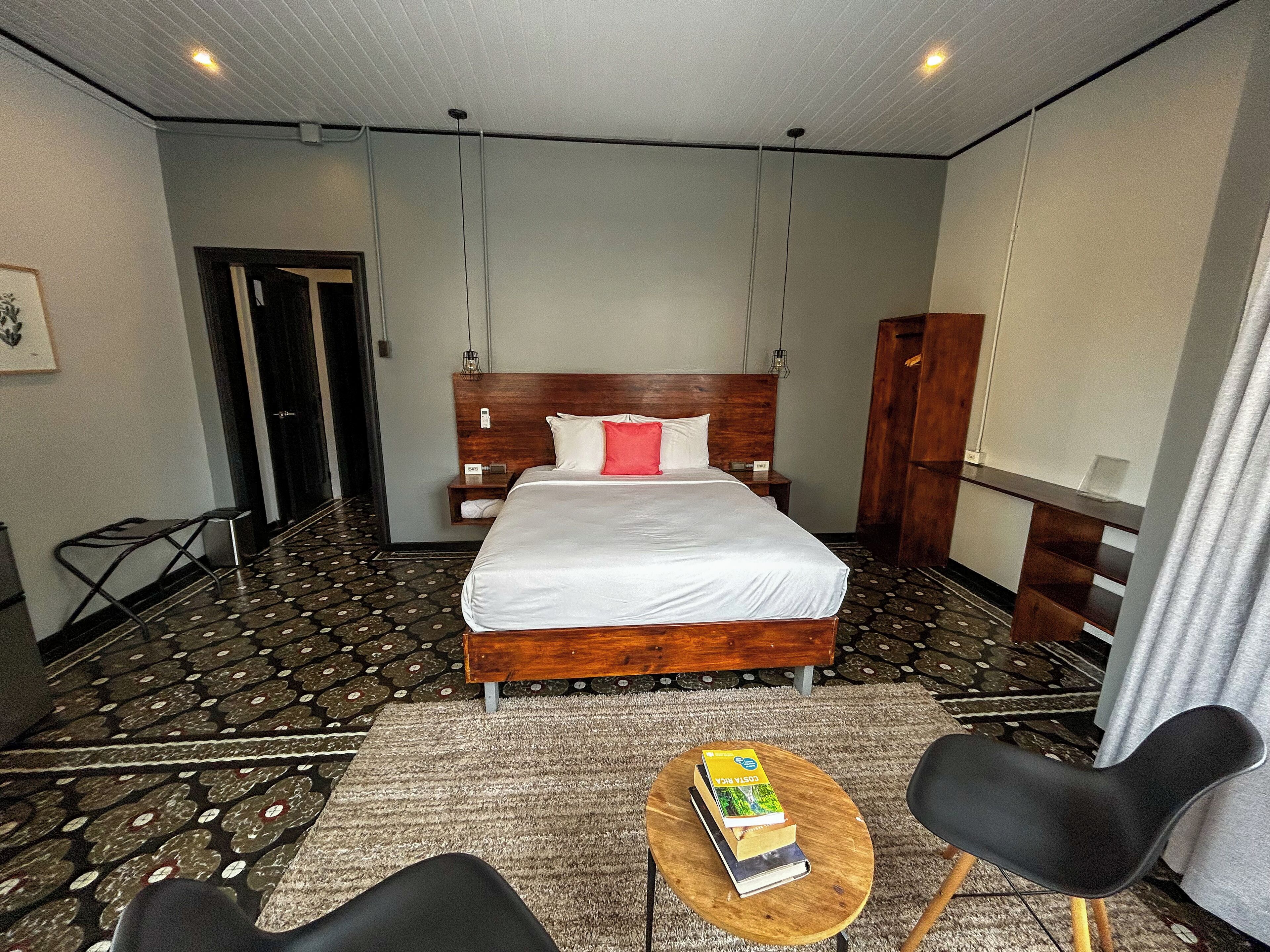 A hotel room with a large bed, a wooden headboard, a small bedside table, a chair, and a round coffee table with books.