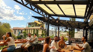 An outdoor café with wooden tables and chairs, a covered seating area, and a view of buildings in the background. - Viajero San José Hostel (San José)