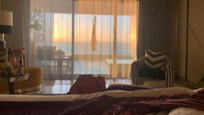 Camera d'albergo con un letto, una sedia, un tavolo con una lampada e vista sul tramonto attraverso ampie finestre.