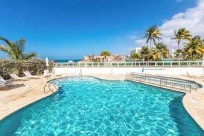 Una piscina con acqua cristallina, circondata da sdraio e palme, con vista su una zona costiera e edifici sullo sfondo.