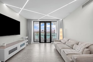 Sala de estar moderna com televisão de tela plana, sofá, mesa de centro e porta de vidro deslizante para uma varanda com vista para a cidade.
