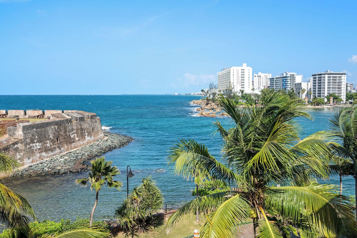 A coastal cityscape with a historic fort, modern high-rise buildings, and palm trees along the shoreline.