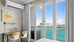 A hotel room with a bed, a desk with two chairs, an air conditioner, and a large window with a city view.