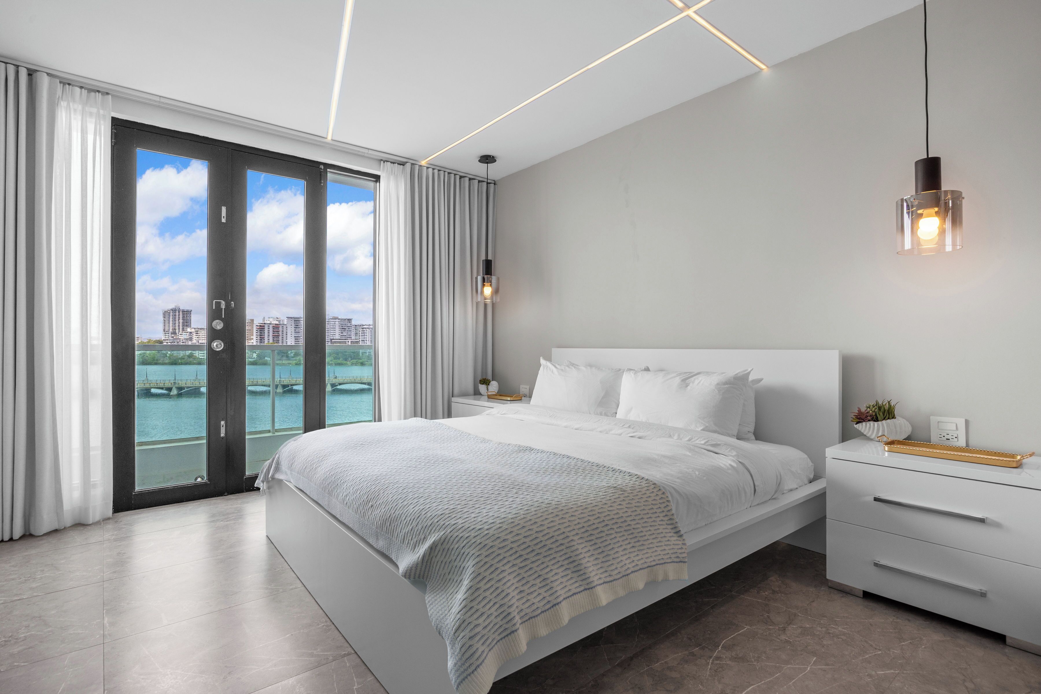 Quarto moderno com uma cama grande, uma mesa de cabeceira, uma luminária pendente e vista para a água através de uma porta de correr.