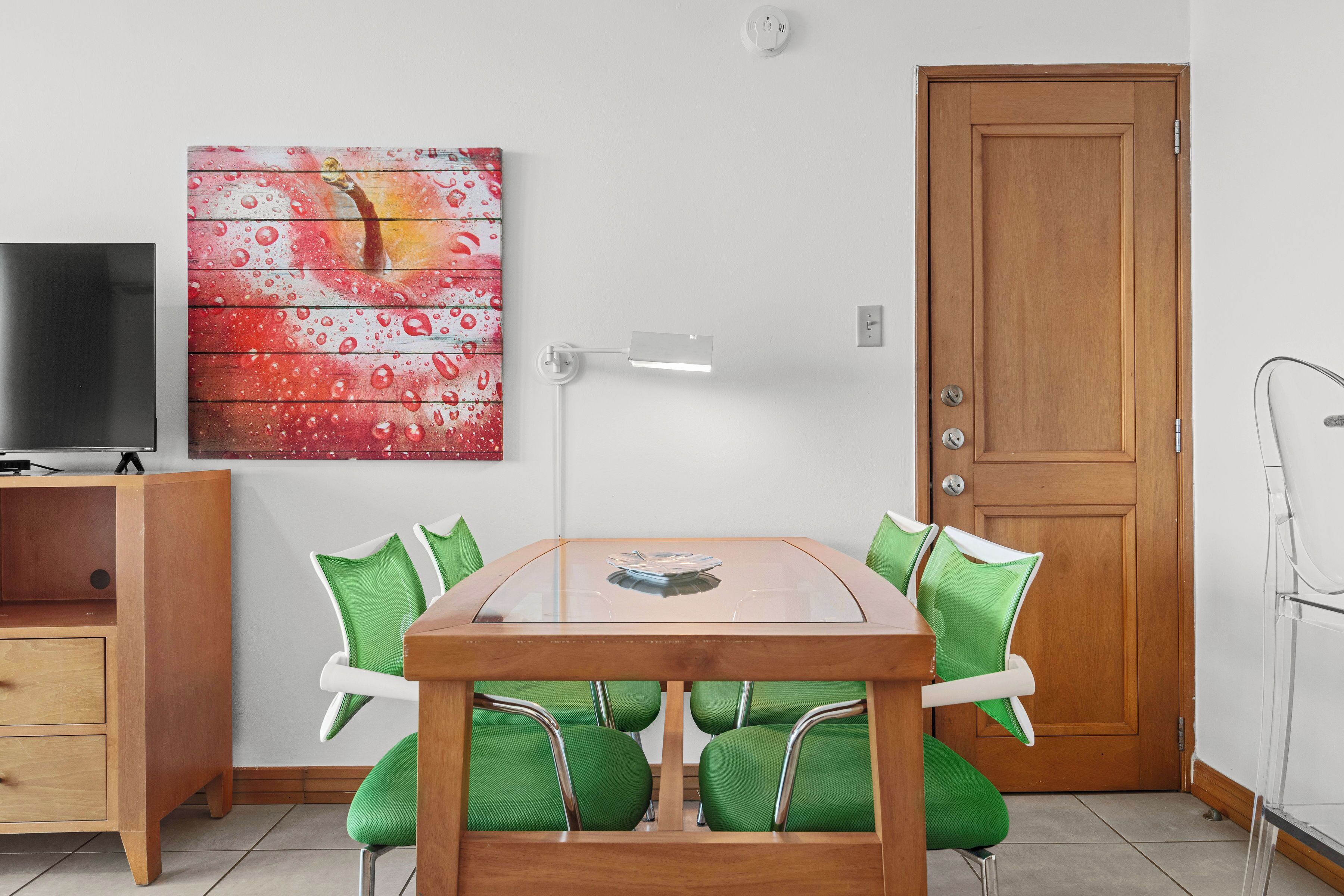 A dining area with a glass-top table, green chairs, a wooden sideboard, and a painting on the wall.