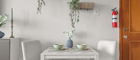 A dining area with a white table, two chairs, a vase with flowers, and hanging plants on the wall.