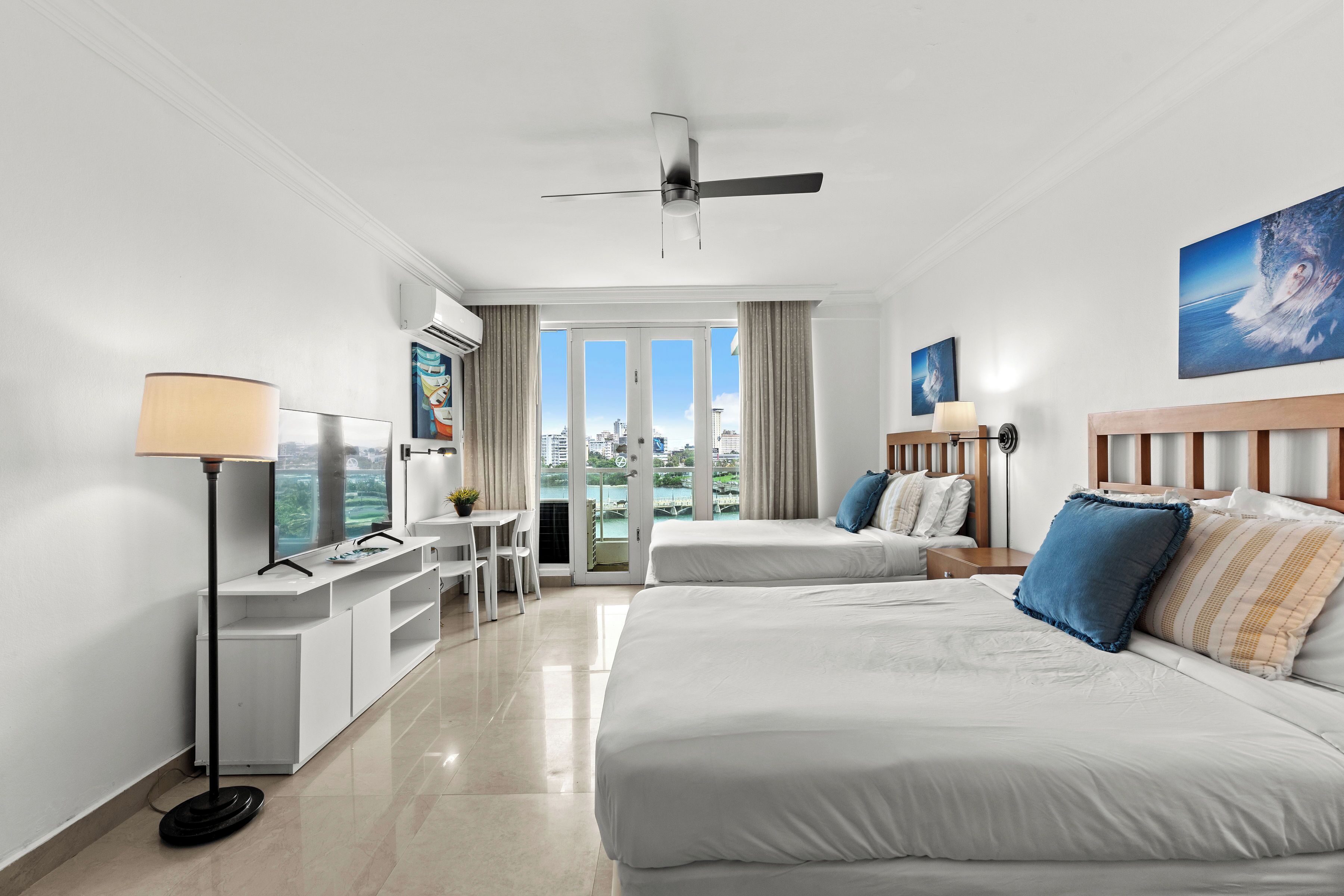 A hotel room with two beds, a desk, a chair, a ceiling fan, and a view of the city through large windows.