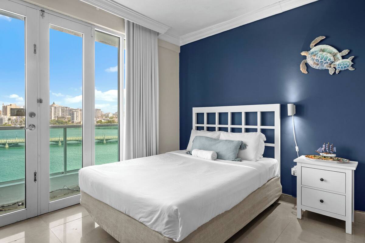 A bedroom with a large bed, a nightstand, a decorative turtle wall art, and a view of the city through a glass door.