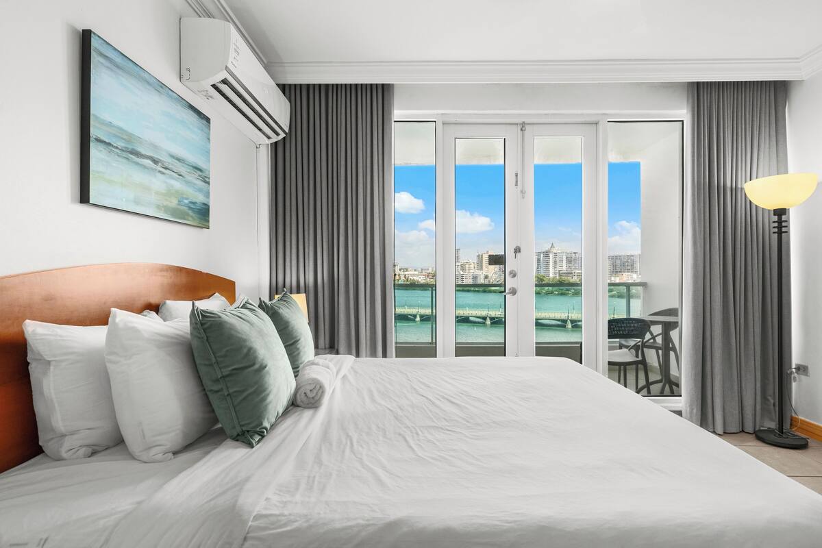 A hotel room with a large bed, a painting on the wall, an air conditioner, and a view of the city through the window.