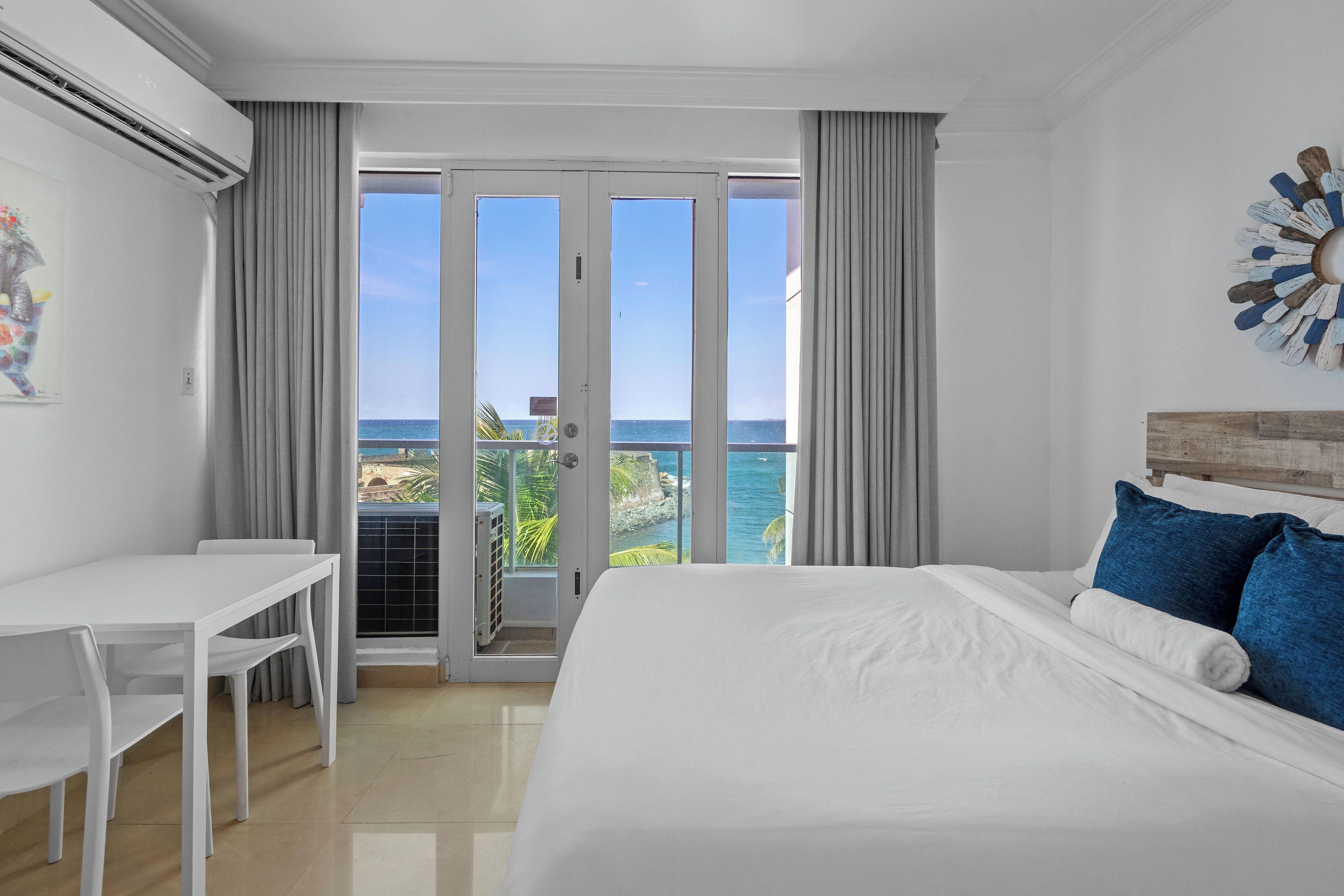 A hotel room with a bed, a desk, and a view of the ocean through a sliding glass door.