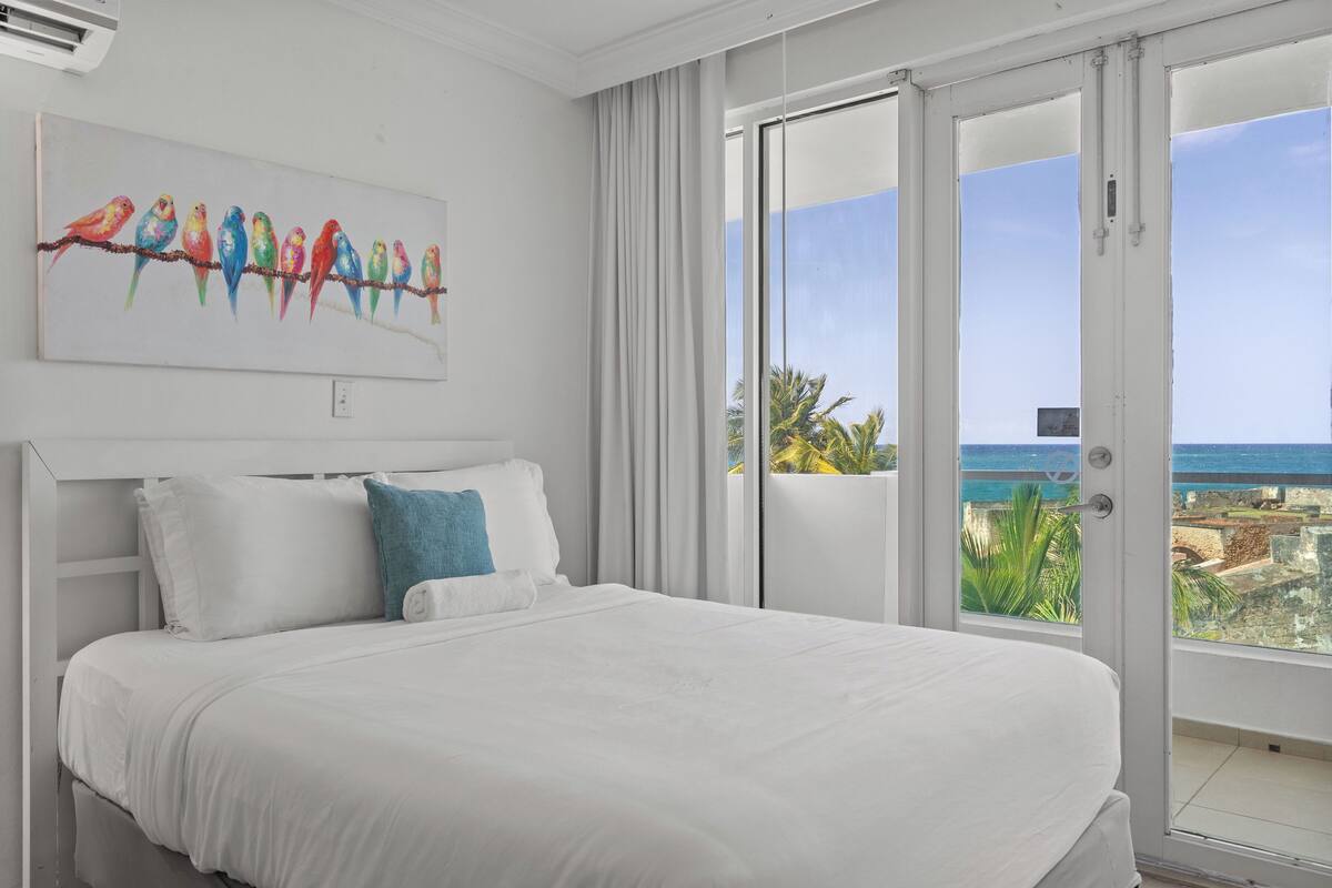 A hotel room with a large bed, a painting of colorful birds, and a view of the ocean.