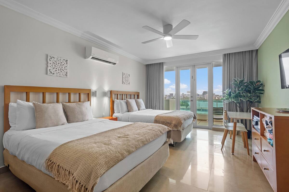 A hotel room with two beds, a ceiling fan, a desk with chairs, and a large window with a city view.
