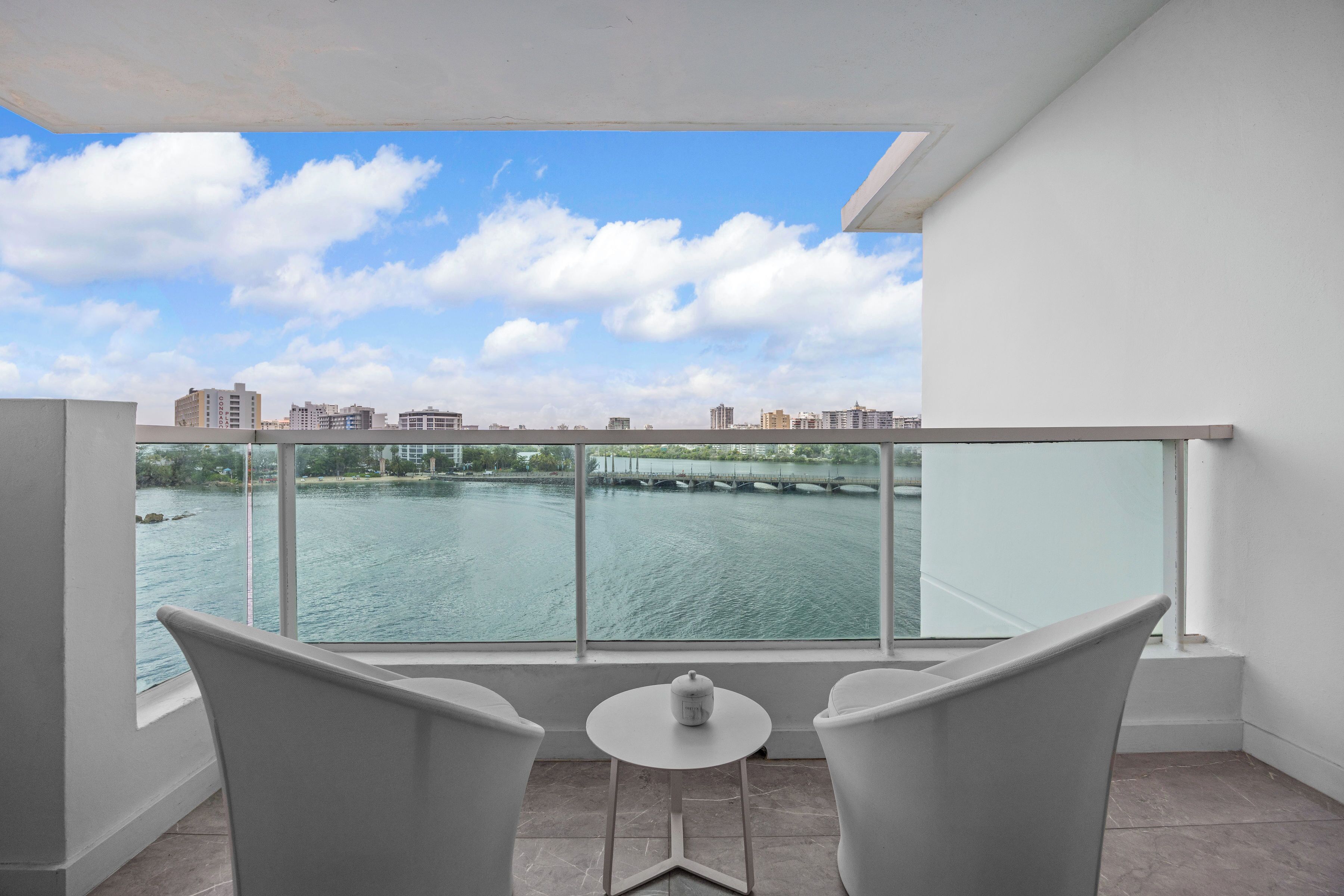 Un balcon avec deux chaises et une petite table, donnant sur une étendue d’eau et sur le paysage urbain.