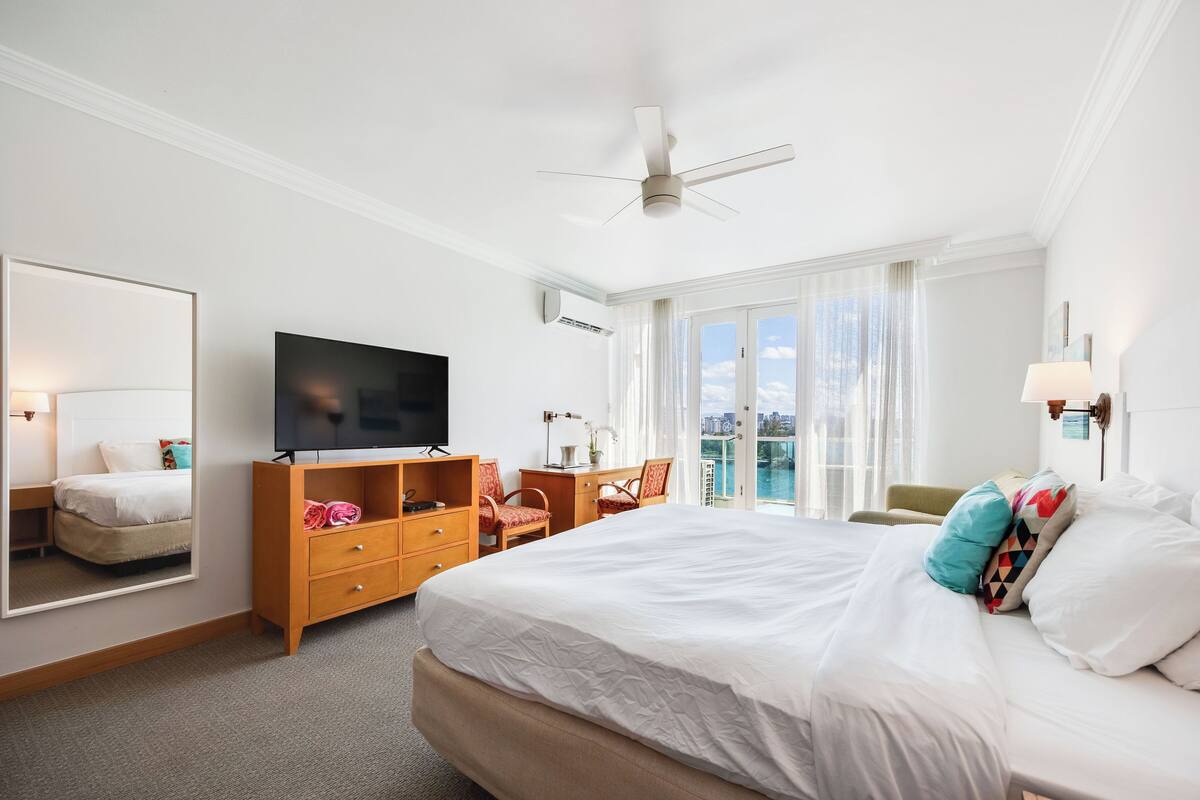 A hotel room with a large bed, a TV, a desk with a chair, a mirror, and a balcony with a view.