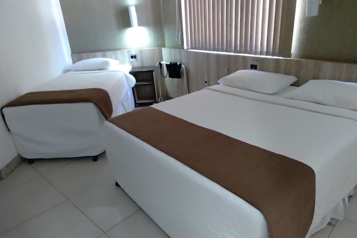 A hotel room with two beds, a bedside table, a lamp, and a window with blinds.