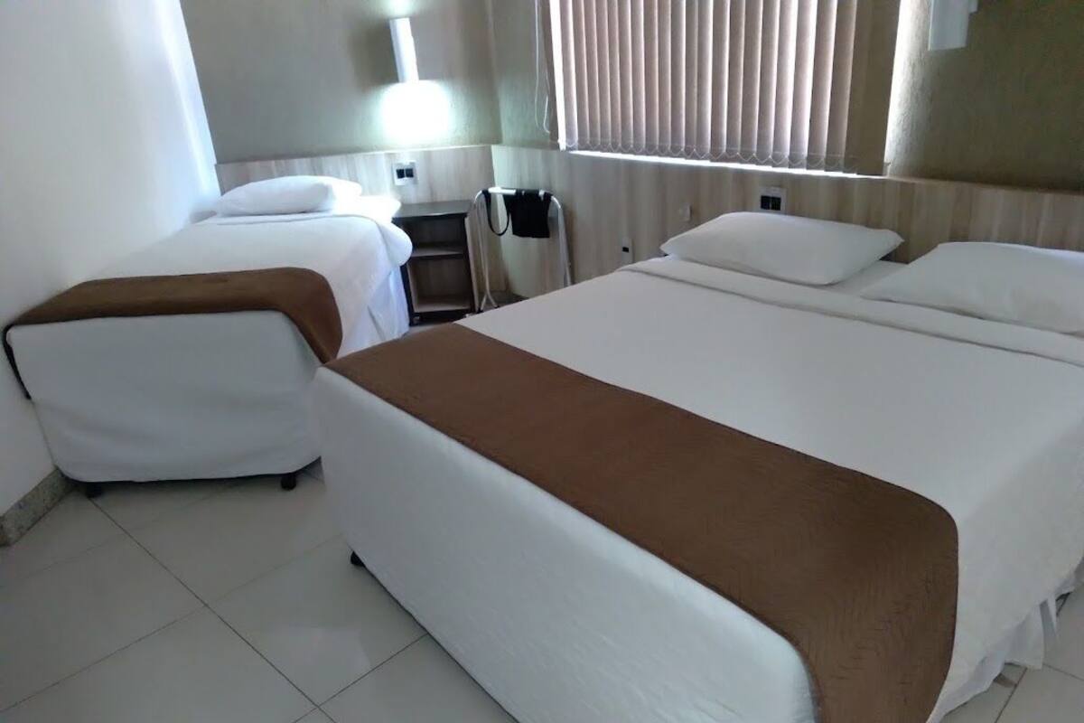A hotel room with two beds, a bedside table, a lamp, and a window with blinds.