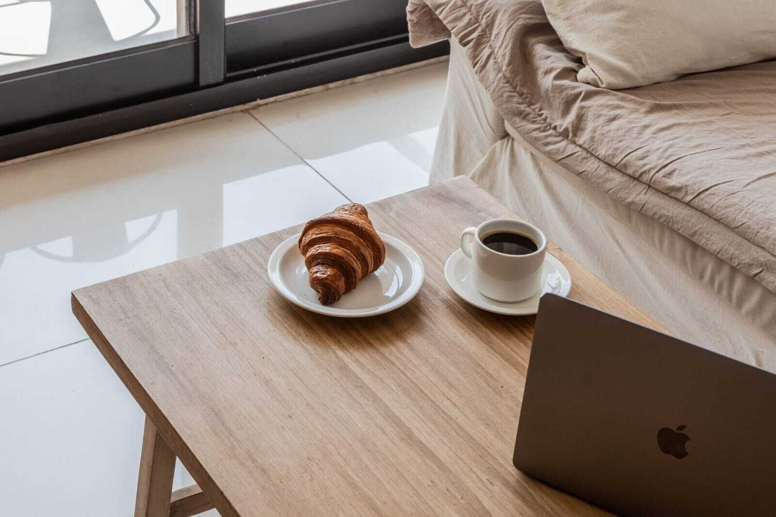 A laptop on a wooden table with a croissant and a cup of coffee beside it.