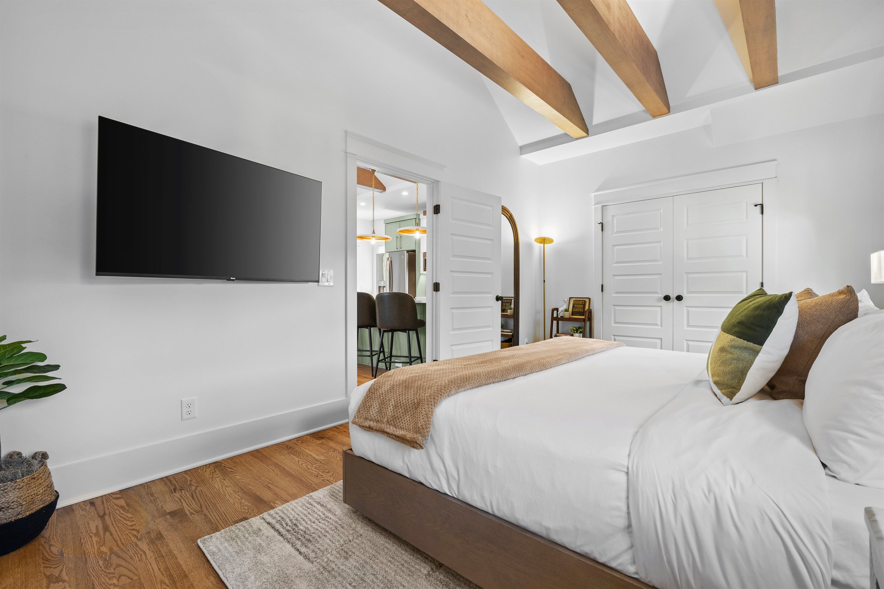 A bedroom with a large bed, a flat-screen TV mounted on the wall, and a wooden ceiling.