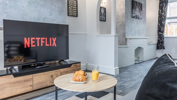 Un salon moderne avec un téléviseur à écran plat affichant le logo « NETFLIX », une table basse en bois sur laquelle se trouve une assiette de nourriture et un verre de jus d’orange, et un canapé noir.