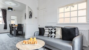 Un salon moderne avec un canapé gris, une table basse ronde en bois sur laquelle sont posés des croissants, et un coin repas avec une table et des chaises.