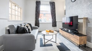Un salon moderne avec un canapé gris, une table basse, un téléviseur posé sur un meuble en bois et une fenêtre avec des rideaux.