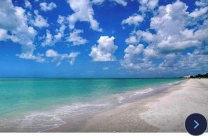 Ein Strand mit klarem türkisfarbenem Wasser und weißem Sand, unter einer teils bewölkten Himmel.