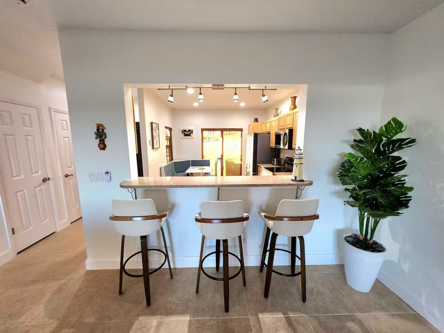 A kitchen with a bar area, three bar stools, a potted plant, and a view into the dining area.