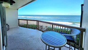A covered balcony with a view of the ocean, a round table with a blue cushion, and a wicker chair. - Sea Breeze Bungalow (Melbourne Beach)
