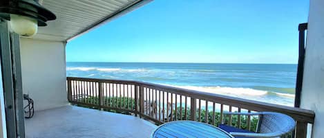 A covered balcony with a view of the ocean, a round table with a blue cushion, and a wicker chair.