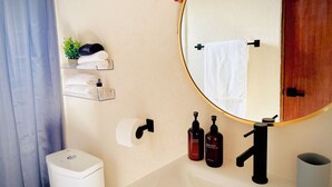 Une salle de bain avec un miroir rond, un lavabo blanc, un robinet noir, des toilettes et une étagère avec des serviettes et des articles de toilette.