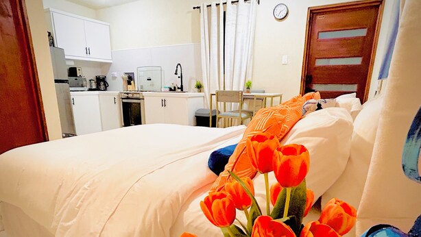 Une chambre d’hôtel avec un lit, une petite table, une chaise, un coin cuisine et une horloge au mur.