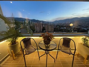 Un balcon avec deux fauteuils en osier et une petite table, d’où l’on peut admirer un panorama sur la ville, avec ses bâtiments et ses montagnes.