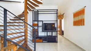 Ein modernes Interieur mit einer Holztreppe, einem schwarzen Metallgeländer, einem Fernseher auf einem schwarzen Sockel und einer Hängelampe.