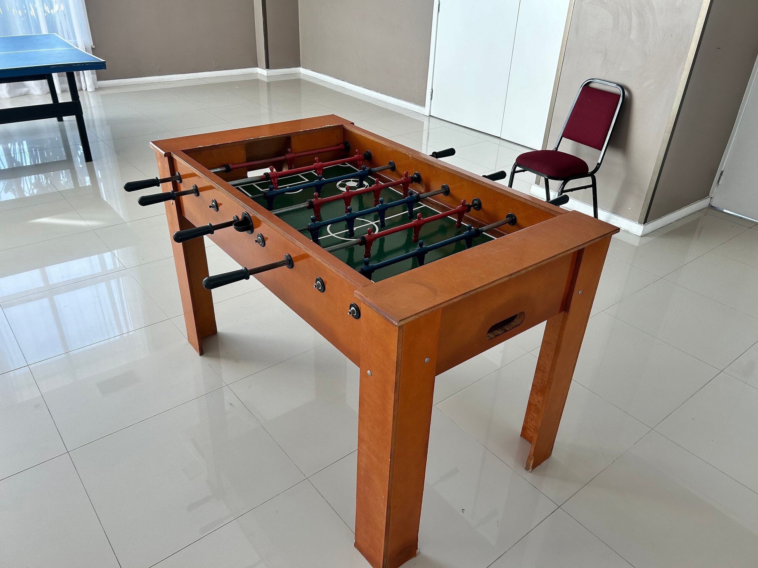 A foosball table in a room with a tiled floor, a red chair, and a table in the background.