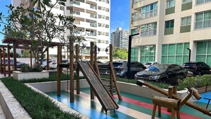 Un parque infantil con un tobogán y estructuras para escalar, rodeado de vegetación y un estacionamiento con coches.