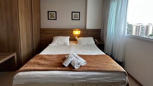 Una habitación de hotel con una cama grande, un cabecero de madera, dos cuadros enmarcados en la pared, una lámpara de noche y una ventana con cortinas.