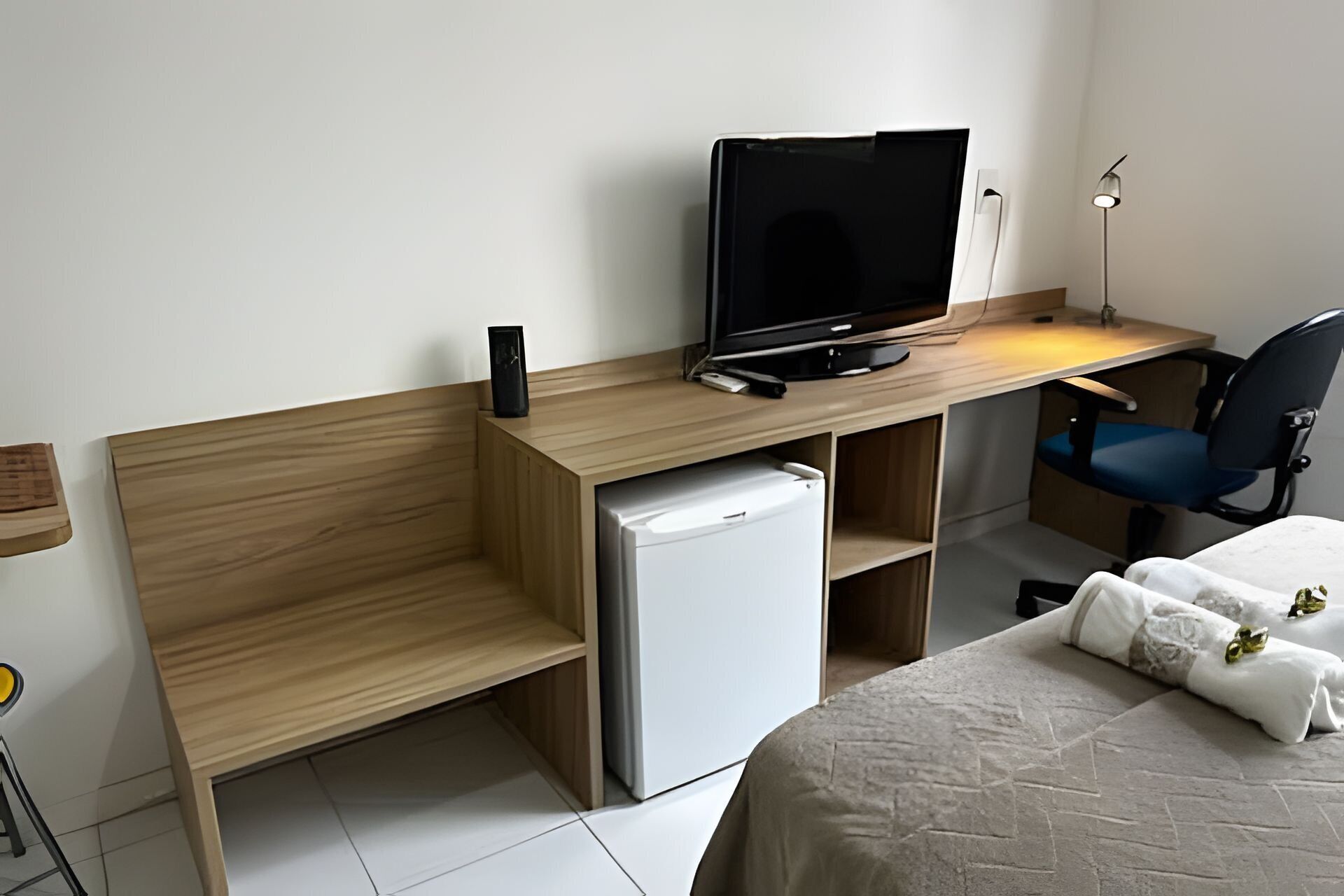 A compact hotel room with a wooden desk, a television, a small refrigerator, a single chair, and a bed with a white cover.