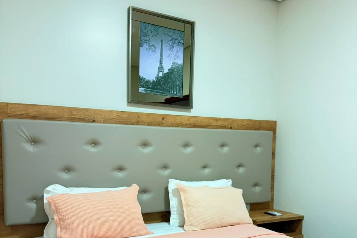 A bedroom with a tufted headboard, two pillows, a bedspread, a nightstand, and a framed picture of the Eiffel Tower.