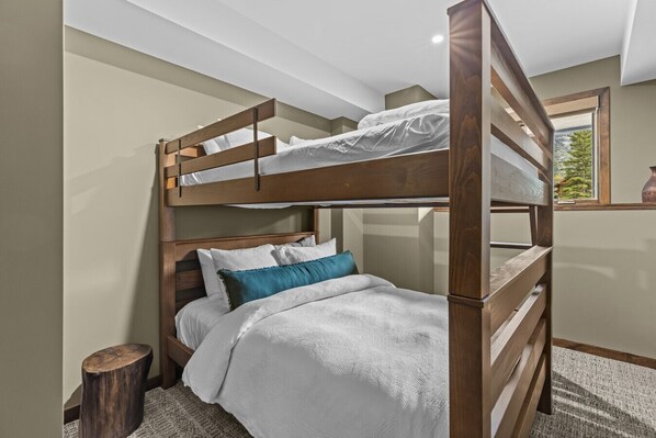 A bunk bed room with a wooden headboard, a small wooden stool, and a window with a view of trees.