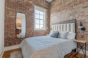 Une chambre avec un mur en briques, une grande fenêtre, un lit avec une tête de lit, un miroir et une table de chevet avec une lampe.