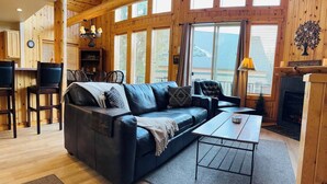 A cozy living room with a black leather sofa, a wooden coffee table, and a fireplace. There are large windows allowing natural light and a view of the outside. - Creekside Chalet with private hot tub (Redmond)