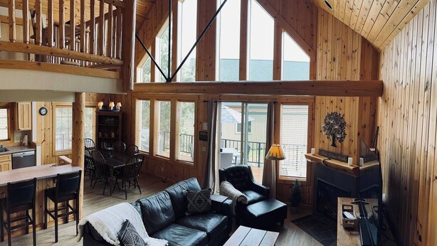 A spacious living area with a high ceiling, wooden beams, a fireplace, and large windows offering views of the outdoors. - Creekside Chalet with private hot tub (Redmond)